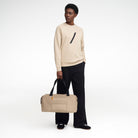 A female model stands at angle to camera, wearing the taupe coloured Volvo Duffle Bag. We see shoulder and cross-body straps, water-repellent exterior and Volvo Diagonal logo.