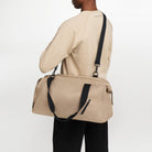 A female model stands at angle with back to camera, wearing the taupe coloured Volvo Duffle Bag. We see shoulder and cross-body straps, water-repellent exterior and Volvo Diagonal logo.