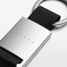 A close-up view of the Volvo Key Chain in black.  We see the solid metal with Volvo logo at the centre, with strap and key ring at the top.