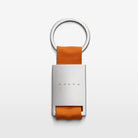 The Volvo Key Chain in orange against a white background. We see the solid metal with Volvo logo at the centre, with strap and key ring at the top.