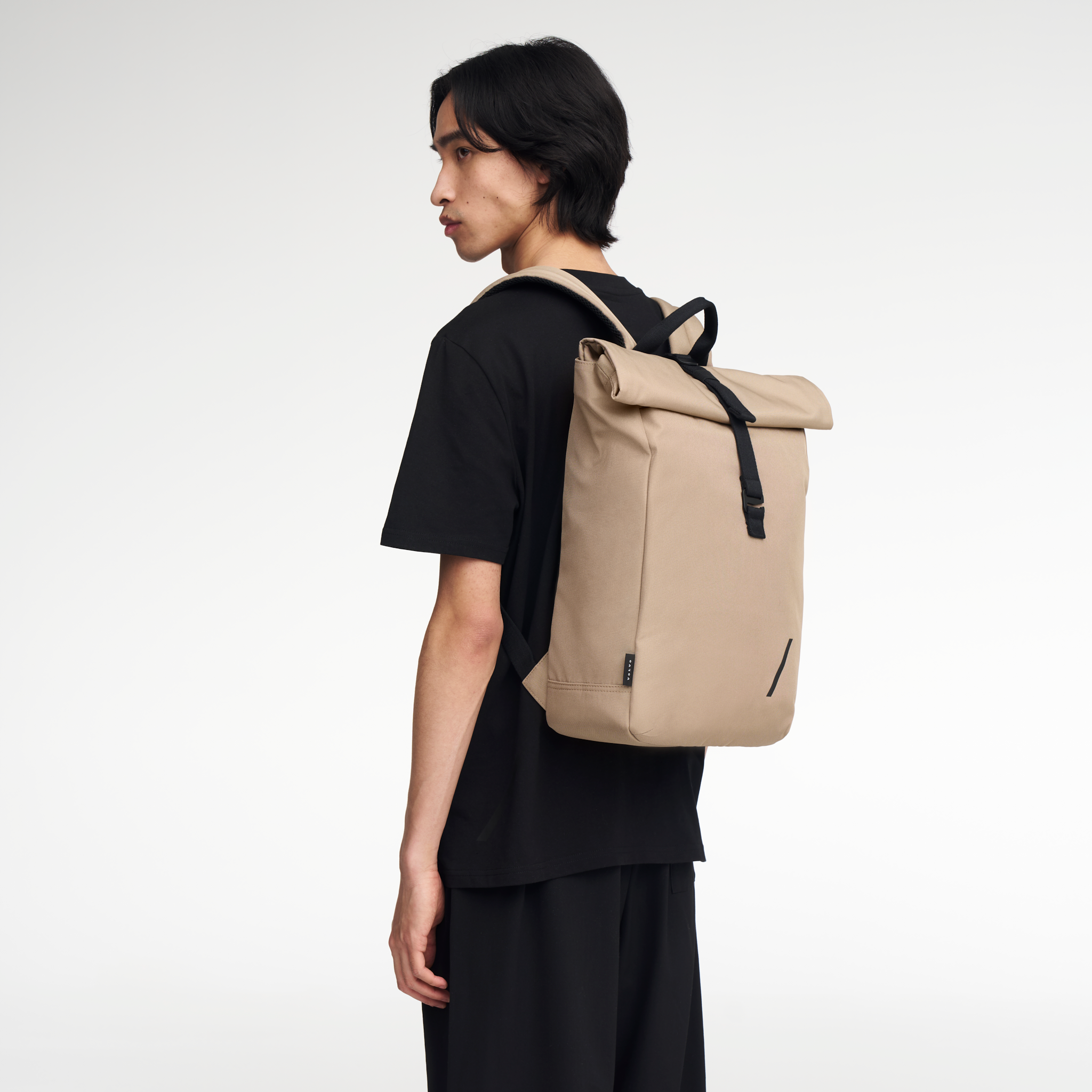 Male model stands at angle with back to camera, wearing the taupe coloured Volvo Backpack. Adjustable straps and Volvo Diagonal logo are visible.